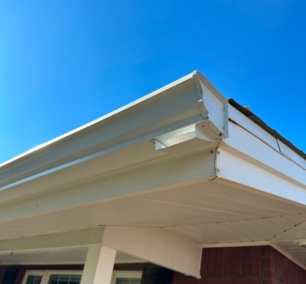 Newly installed white seamless aluminum rain gutter on the roof edge of a brick house against a clear blue sky.